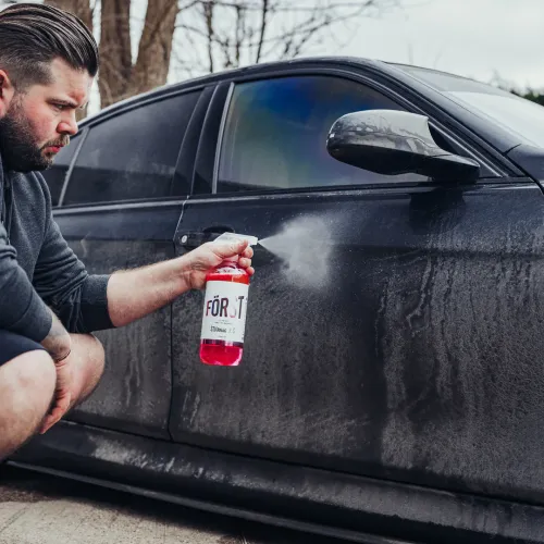 Man sprays cleaning solution from a red and white bottle labeled "FÖRST" onto the dirty side of a parked black car outdoors on a cloudy day with bare trees in the background.