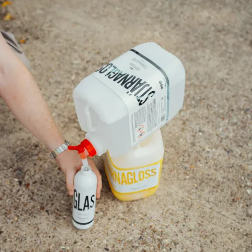 A person refills a small white bottle labeled "GLASS" from a larger white container labeled "STRAMAGLASS," placed upside down on a yellow container labeled "NAGLOSS," in an outdoor setting with a gravel ground.