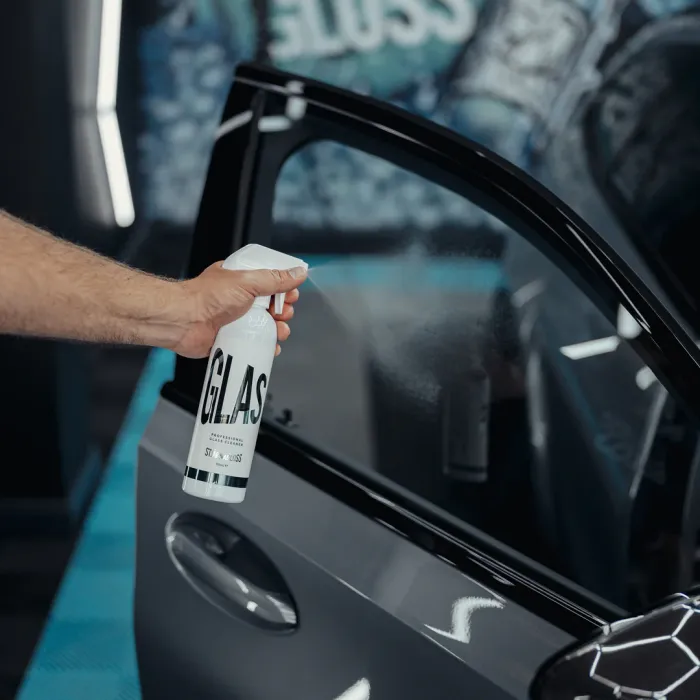A hand sprays glass cleaner from a bottle labeled "GLAS" onto a car window in a garage with blue and white equipment in the background.