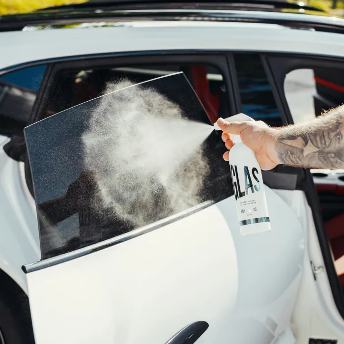 A tattooed arm sprays cleaner labeled "GLAS" onto a car's side window beside an open white car door on a sunny day.