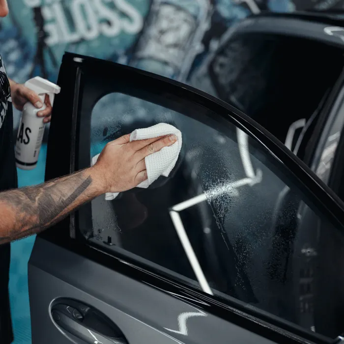 A hand holding a towel cleans a car's side window with a spray bottle in a workshop, with graffiti art on the walls in the background.
