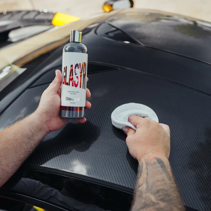 A person holds a bottle labeled "GLASIR PROTECTION GLAZE by STJÄRNAGLANS" while another hand applies a product with a circular pad onto a glossy, dark car surface outdoors.