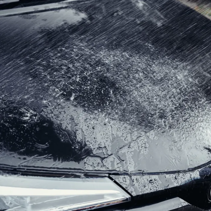 Rainwater splashes on a shiny black car hood, creating wet streaks and droplets; a part of the headlight is visible, emphasizing an outdoor scene.