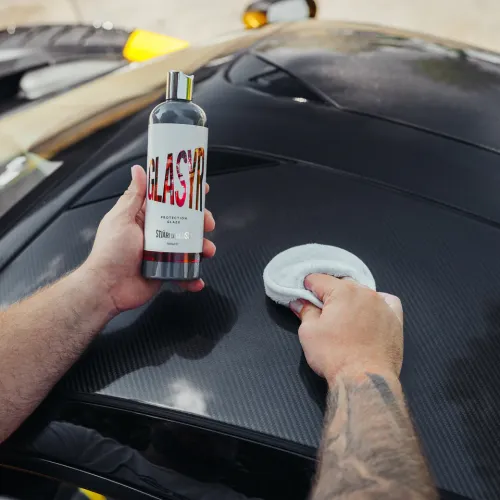 Hands hold a bottle labeled "GLASIR Protection Glaze" and a microfiber pad near a black car’s carbon fiber hood, ready to apply the product for protection.