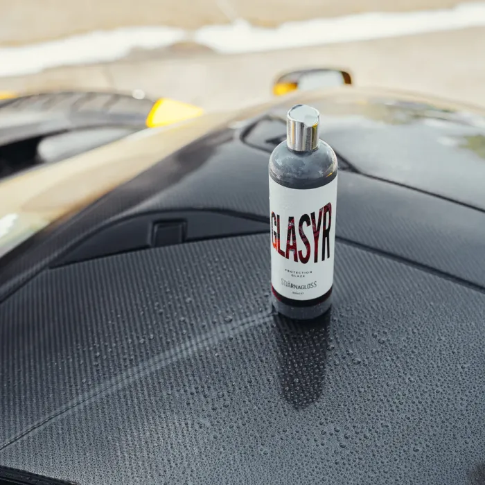 A bottle labeled "GLASYR Protection Glaze by STJARNAGLOSS" is resting on the wet, sleek surface of a black car roof, with blurred street and sidewalk in the background.