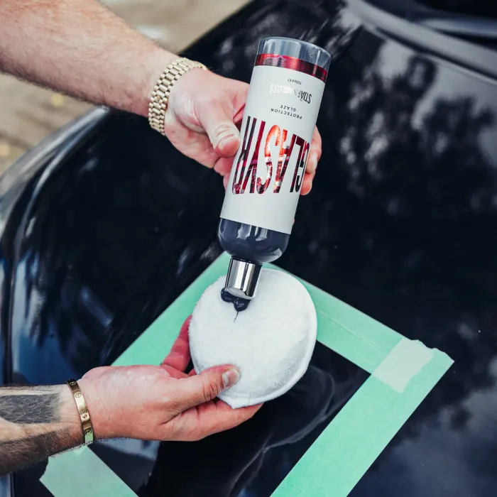 A person applies "STAR LABS CSLAYS PROTECTION GLAZE" from a bottle onto a white applicator pad, over a black car's surface marked with green tape.