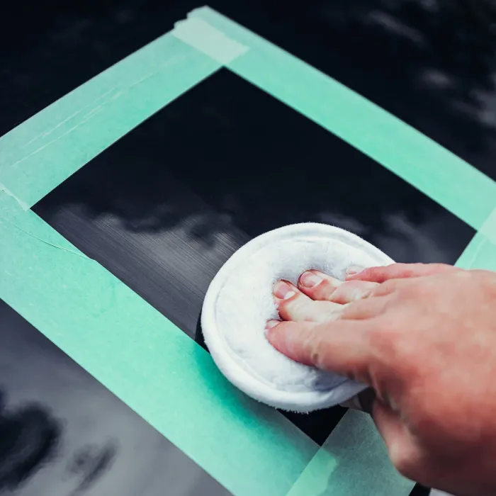 A hand polishes a taped-off section of a car's black surface using a white cloth pad in an outdoor setting.