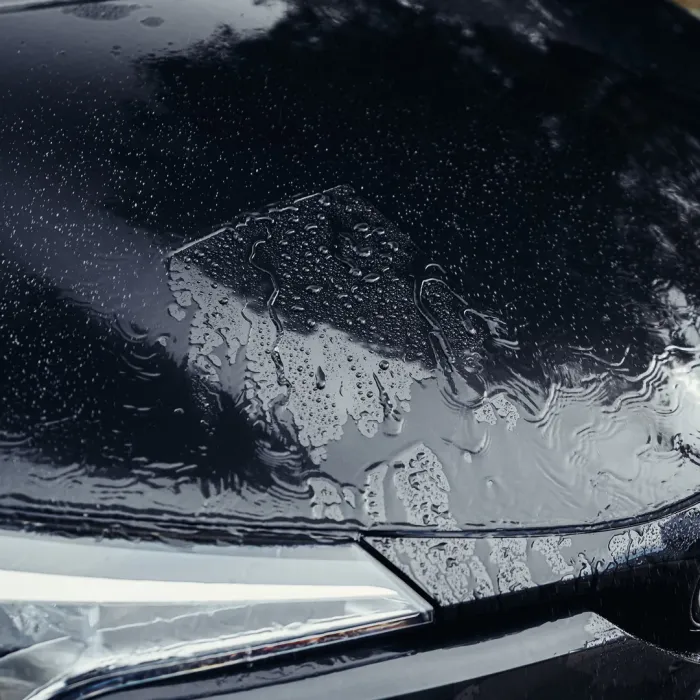 Raindrops rest on the black, glossy surface of a car hood, highlighting the vehicle's clean lines and modern headlight design, with reflections of trees and a cloudy sky overhead.