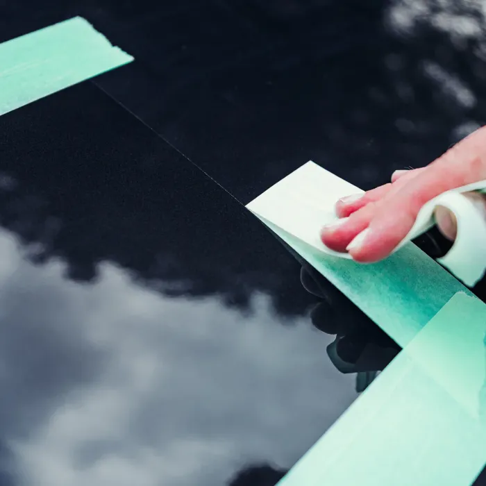 A hand is applying green masking tape to a glossy black surface with a faint reflection of clouds visible.