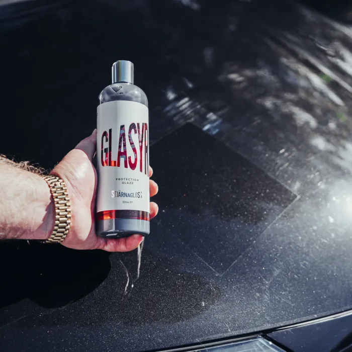 A hand holds a bottle of "Stjärnagloss Glasyrl Protection Glaze" in front of a black car hood reflecting light and surface textures in an outdoor setting.