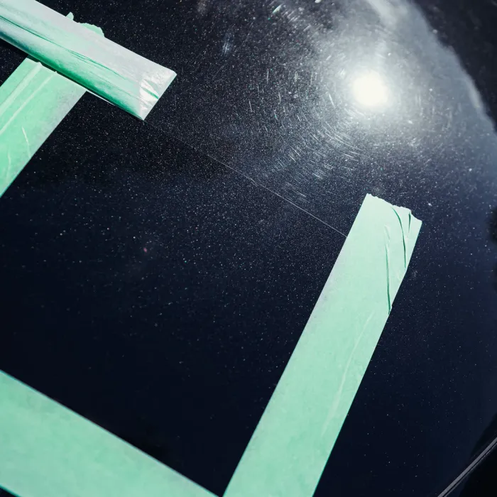 Green painter's tape forms a large rectangle on a dark, glossy surface, possibly a car hood, reflecting sunlight and showing fine scratches.