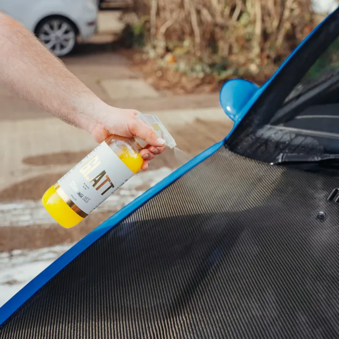 A hand sprays a bottle labeled "GLATT Super Gloss" onto the carbon-fiber hood of a blue car in an outdoor setting, with a blurred white car and foliage in the background.