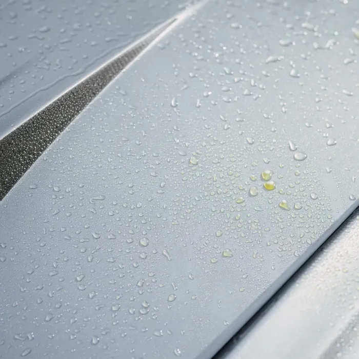 Raindrops are scattered on the silver surface of a car hood, emphasizing a sleek, metallic texture and partially separated by narrow grooves.