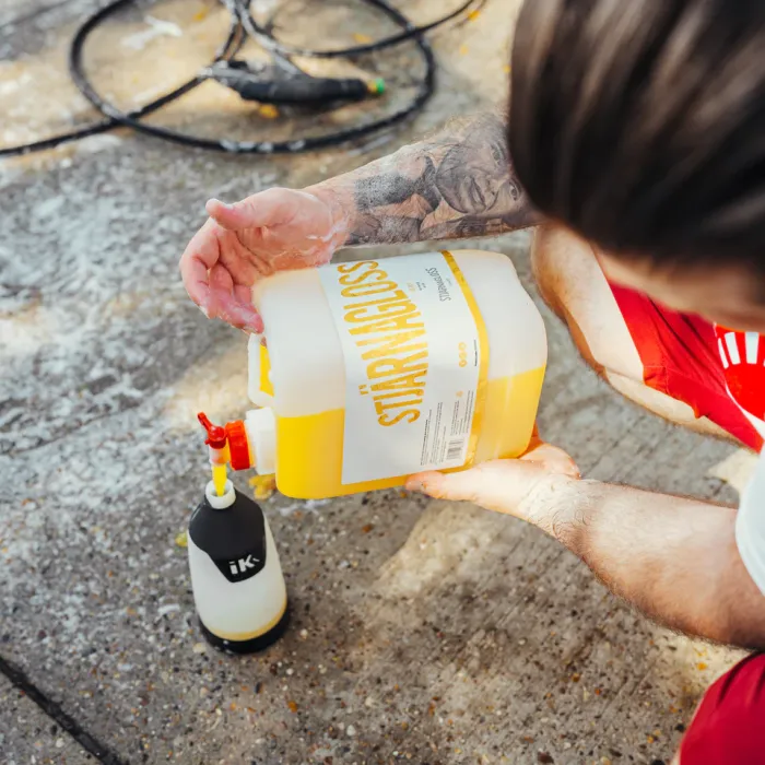 A person in red shorts pours yellow liquid from a "Stjarnagloss" container into a spray bottle on a concrete surface, with a coiled hose and soap suds nearby.