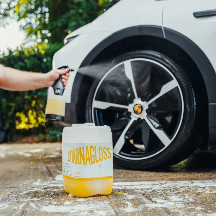 A hand sprays cleaning solution onto a white car's black alloy wheel. "STJÄRNAGLOSS" is written in yellow on a container in the foreground. Context is outdoors with greenery in the background.