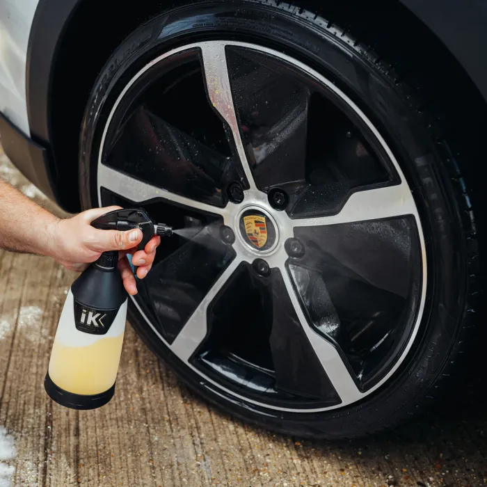 Hand holding an "iK" branded spray bottle, cleaning a black and silver car wheel with a Porsche emblem, on a concrete surface.