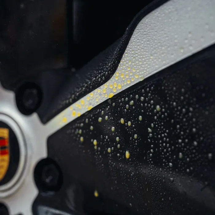 Close-up of a car wheel's black and silver surface with a logo, covered in yellow-colored water droplets, creating a textured appearance, possibly after rain.