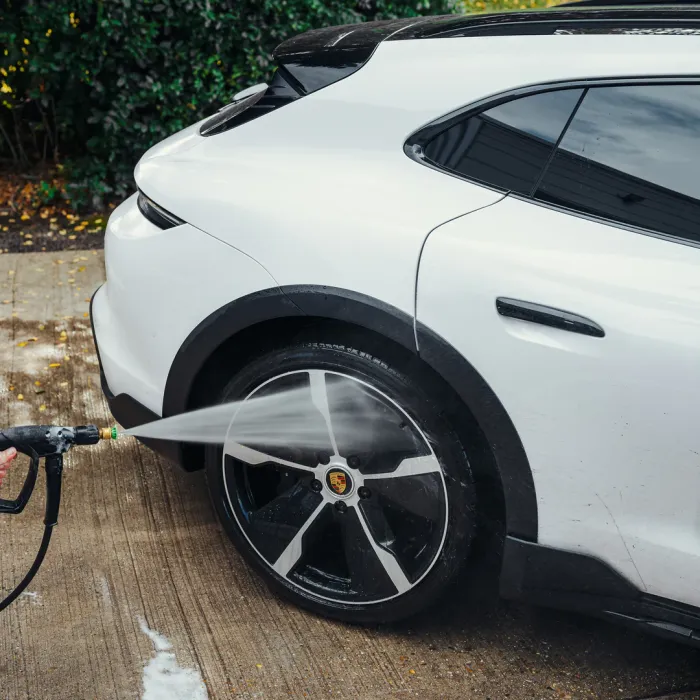 A person power-washes the rear wheel of a white Porsche car on a concrete driveway, surrounded by leafy green bushes.