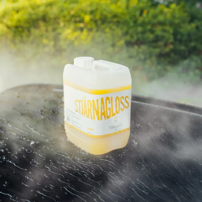 A white and yellow container of "STJÄRNAGLOSS" cleaning solution is placed on a wet, shiny surface, surrounded by mist, with a blurry green hedge in the background.