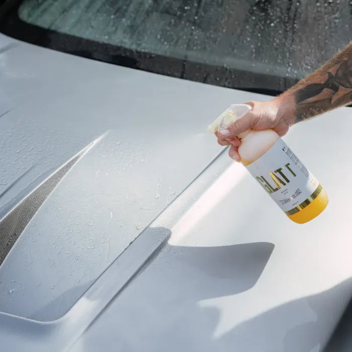 A tattooed arm sprays "GLATT by Stjärnagloss" from a yellow bottle onto the wet hood of a car, emphasizing cleaning or detailing in an outdoor setting.