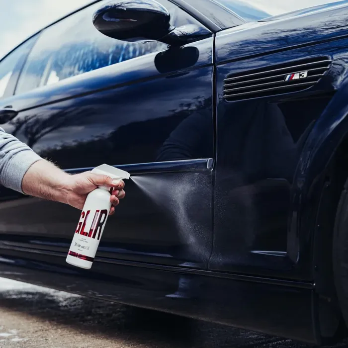 A hand sprays a cleaning product labeled "GLR" onto the side of a shiny dark blue car, parked outdoors near a tree-lined area under a cloudy sky.