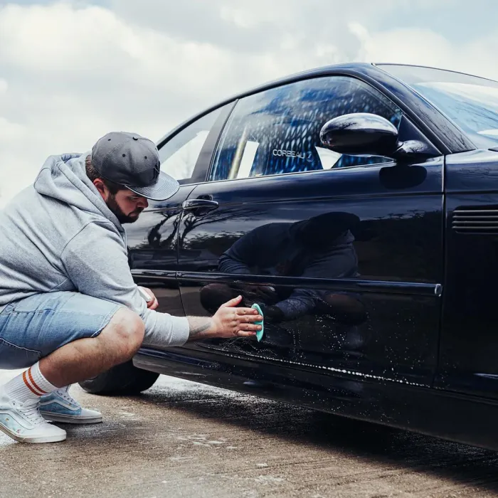 A person in a gray hoodie and cap is crouching and cleaning the side of a black car with a sponge, in an outdoor setting with a cloudy sky.
