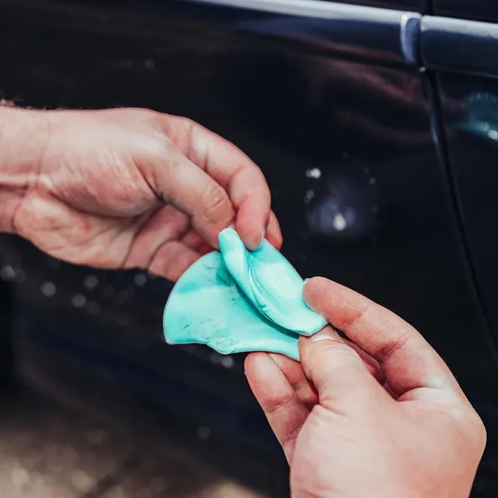 Hands manipulate a blue cleaning putty against a dark car surface outdoors, suggesting vehicle detailing.