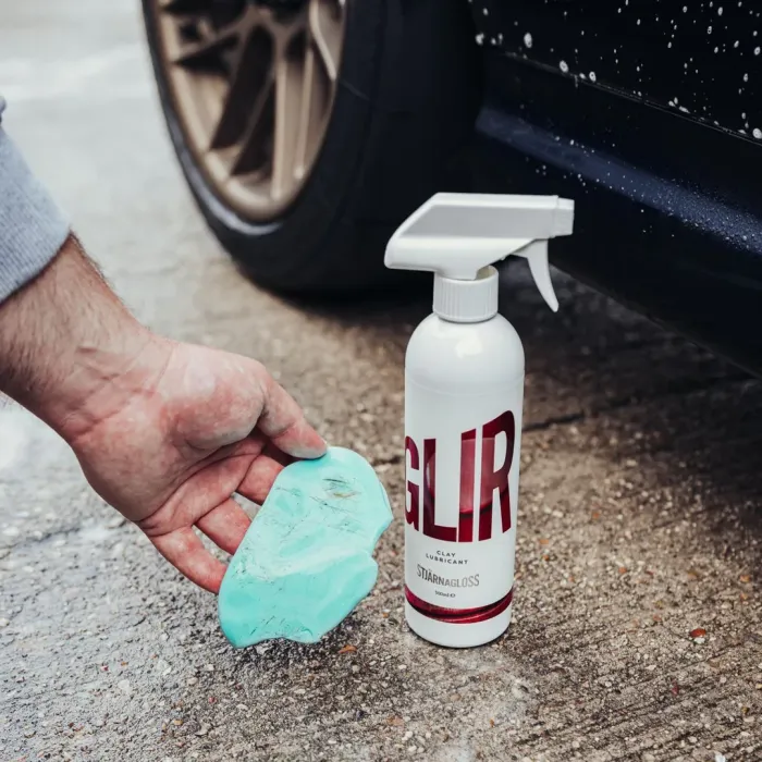 A hand holding a piece of blue clay next to a white spray bottle labeled "GLIR CLAY LUBRICANT STJARNAGLOSS" on wet pavement near a black car's wheel and fender.