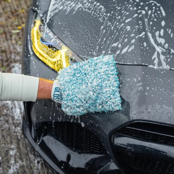 A hand wearing a blue and white cleaning glove labeled "CLEAN GLOSS" scrubs the soapy front of a grey car outside on a wet surface.