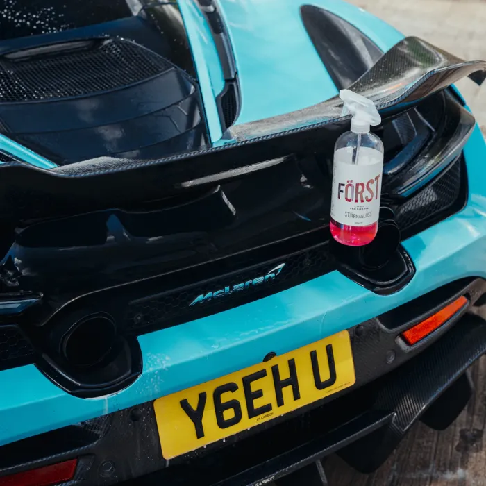 A blue McLaren car with yellow license plate "Y6EH U" holds a spray bottle of "FÖRST SD BANANA GLOSS" on its rear spoiler in an outdoor setting.