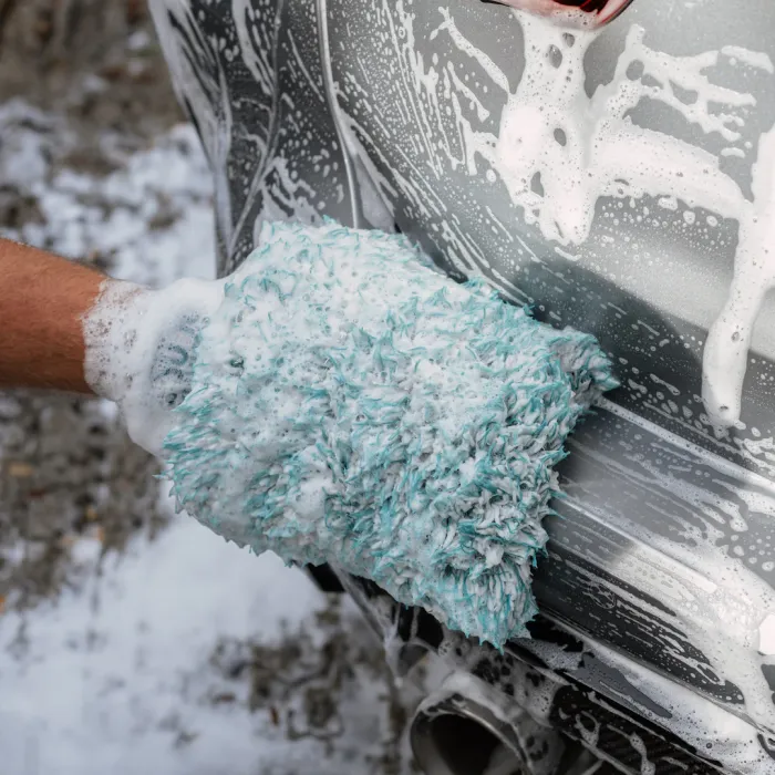 A gloved hand washes a soapy car exterior with a blue and white microfiber mitt, surrounded by suds and wet ground.