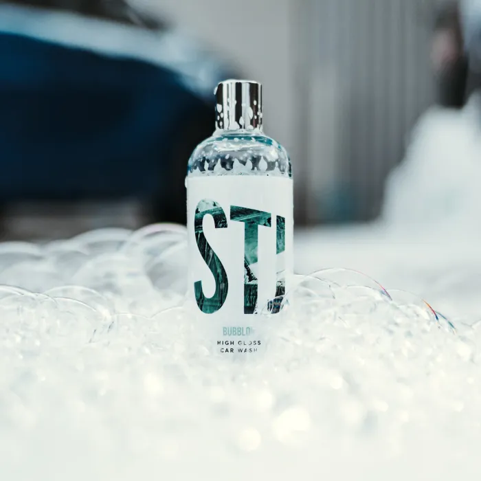 A bottle labeled "STI BUBBLOX HIGH GLOSS CAR WASH" stands among soap bubbles, with a blurred car and urban background.