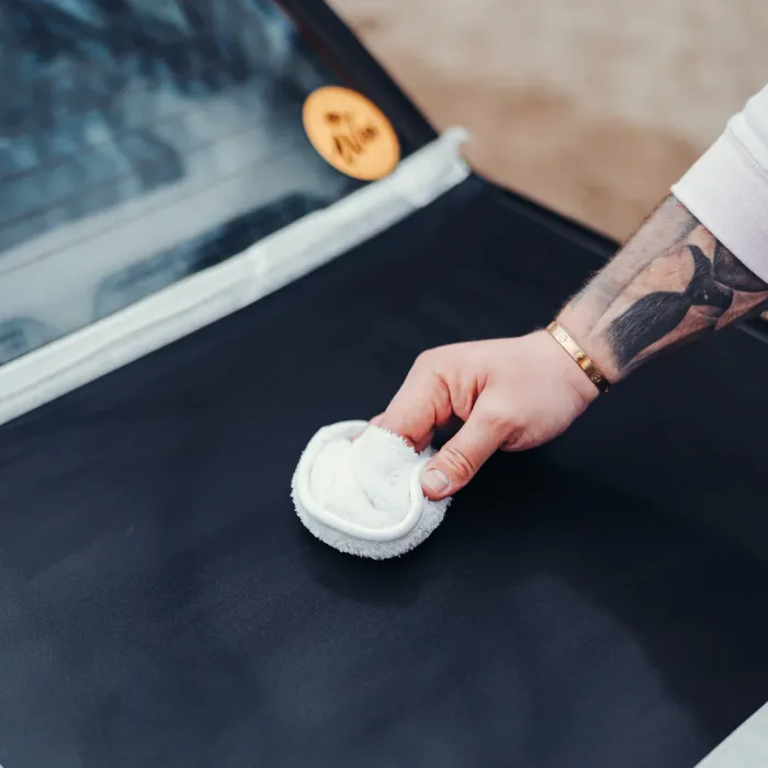 A tattooed arm holds a white cloth pad, applying it to a black surface, likely a car, with a blurred window and an orange circular sticker in the background.