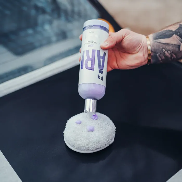 A hand squeezes a bottle of purple-colored polish labeled "ARM" onto a round, white, fluffy buffer pad against a dark surface, possibly a car exterior.