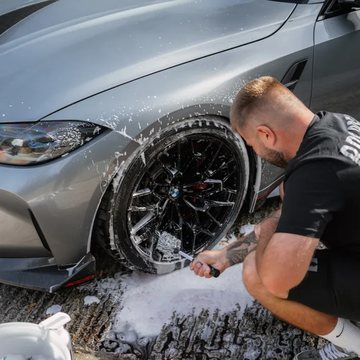A person scrubs the front wheel of a silver BMW car with a brush, creating foam, while crouching on a wet and soapy driveway.