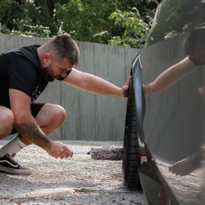 A person squats and cleans a car tire with a brush in an outdoor area, surrounded by a fence and greenery.