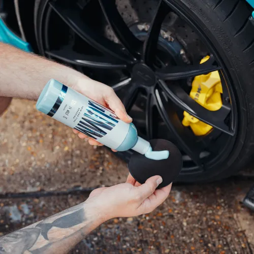A person applies cleaner from a bottle onto a sponge beside a black car wheel with a yellow brake caliper in an outdoor setting.