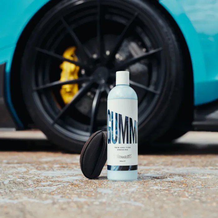 A bottle labeled "GUMMI Trim and Tyre Dressing, Stjärnagloss, 500ml" stands on pavement next to a black applicator pad. In the background, a blue car with black wheels and yellow brake calipers.