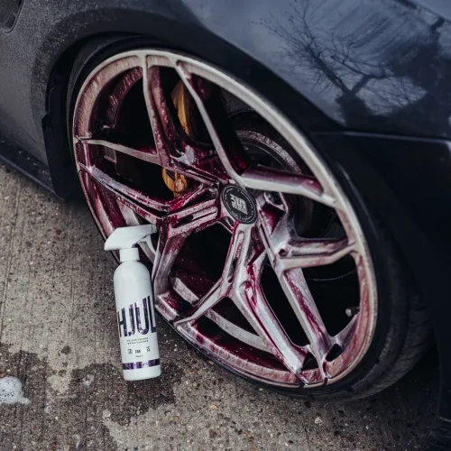 A car wheel with a five-spoke design has a cleaning solution applied, producing a red hue. A white spray bottle labeled "HJUL" rests against the tire on a concrete surface.