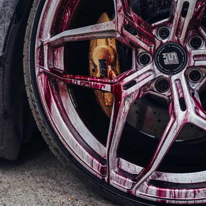 A car tire rim, covered in a red liquid, with visible "WMS" branding, and a yellow brake caliper. The background is part of the car's dirty exterior and a concrete surface.