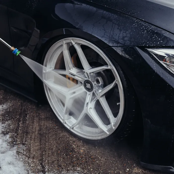 A car wheel being cleaned with a high-pressure washing tool, with water spraying onto the wheel's surface. The vehicle is parked on a wet, textured concrete surface.