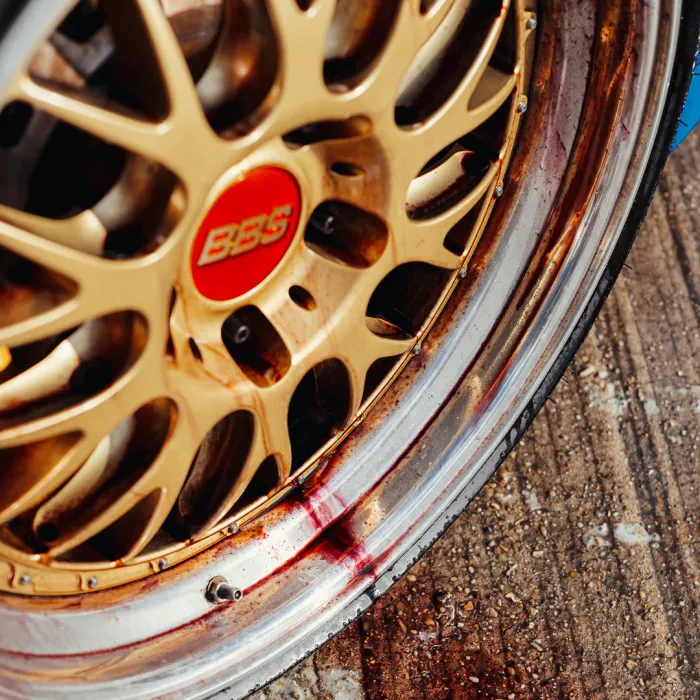 A gold BBS alloy wheel with a red logo has a stained rim, possibly with brake fluid, and is on a wet, textured concrete surface.