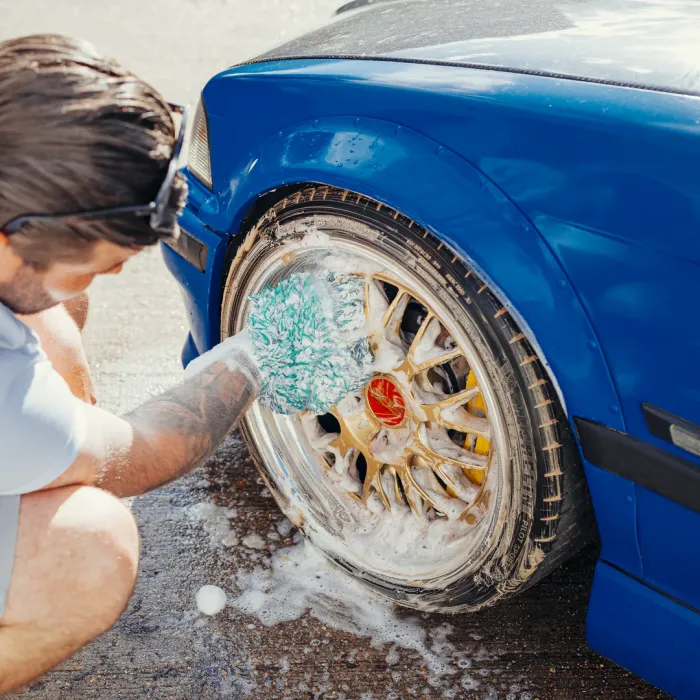 A person kneels, scrubbing the soapy wheel of a blue car with a green glove, on a sunlit pavement.