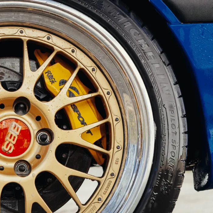 Gold alloy wheel with a red BBS center cap and visible yellow Brembo brake caliper, surrounded by a black Michelin Pilot Sport tire on a wet blue car.