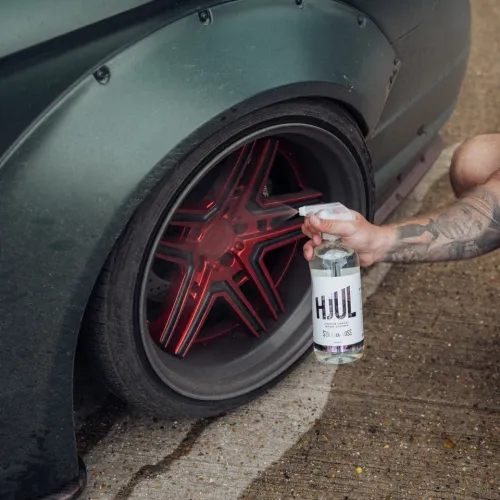 A tattooed arm holding a spray bottle labeled "HJUL" aims it at a car's red, five-spoke wheel rim beside a dark green body panel on a concrete surface.