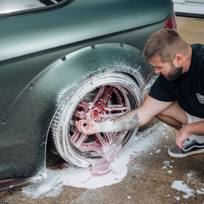 A man kneels and cleans a soapy, deep-dish car wheel, featuring red spokes, in an outdoor setting near a garage.