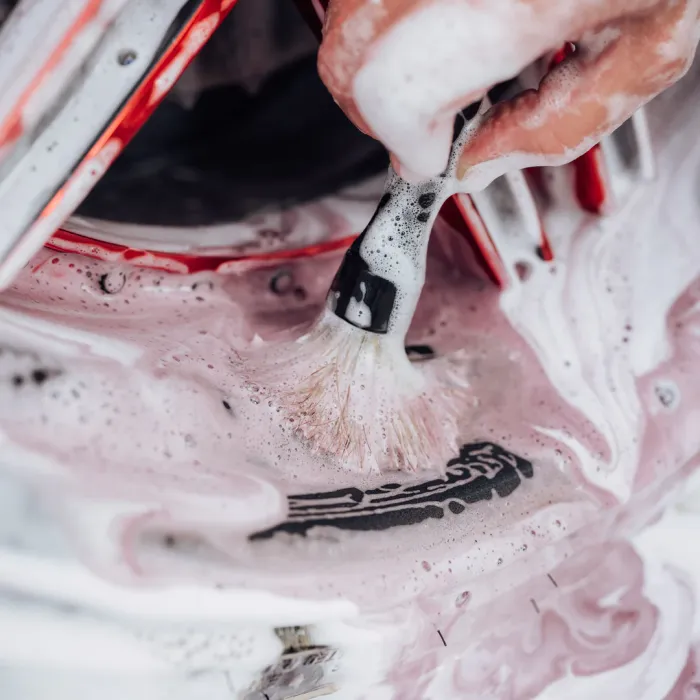 A hand uses a brush to scrub a surface with pink, foamy soap, emphasizing the cleaning action. The surroundings include red- and white-colored objects in a close-up view.