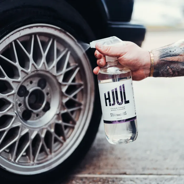 A hand sprays a bottle labeled "HJUL Colour Change Wheel Cleaner" near a car's silver alloy wheel with intricate spokes, set in an outdoor environment with a concrete ground.