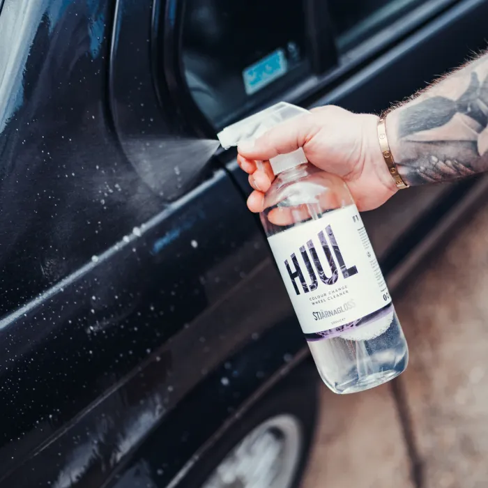 A tattooed arm sprays Hjul Colour Change Wheel Cleanser on a black car door outdoors.