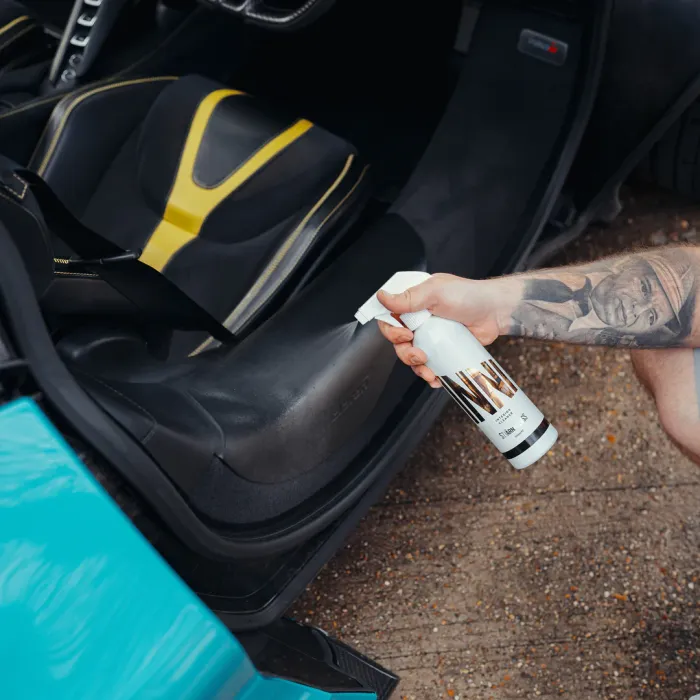 A tattooed hand sprays a car interior with a cleaning product next to a partially opened turquoise car door on a gravelly ground surface.